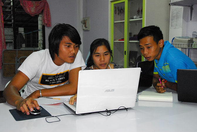 Three young students gather together around a laptop to tune in to an education program.