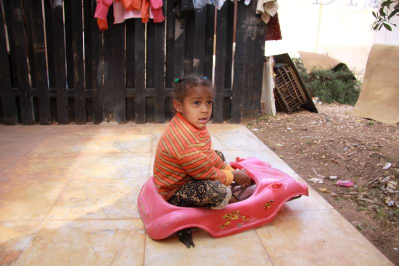بنت صغيرة نازحة تلعب خارج منزلها الجديد في بنغازي، وهو بيت من طابق واحد في مجمَّع شركة سابق. منزل عائلتها جنوبًا في أجدابيا وهو غير ملائم للسكن.