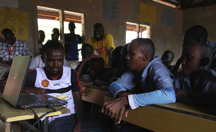 A group of older students gathers in a small, one-room school and circles around a small laptop. 