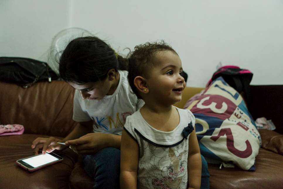 Hanan, 12, checks her phone while one-year-old Yara looks around the family apartment in downtown São Paulo, Brazil. Yara was born at Za'atari refugee camp in Jordan.