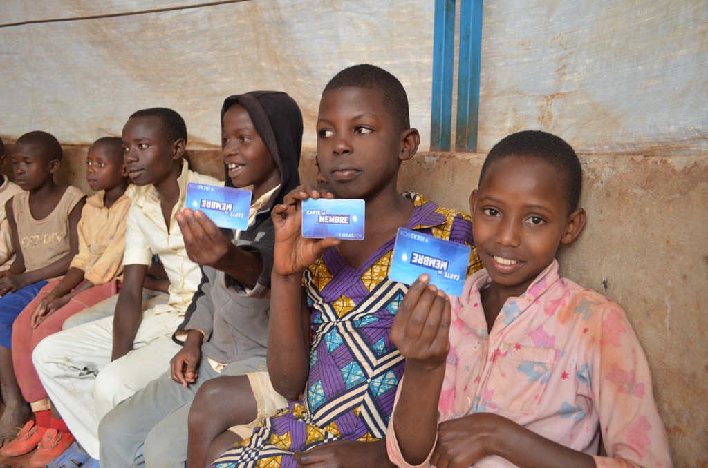 A group of young refugee students sitting on a bench holds up blue library cards for the Ideas Box.