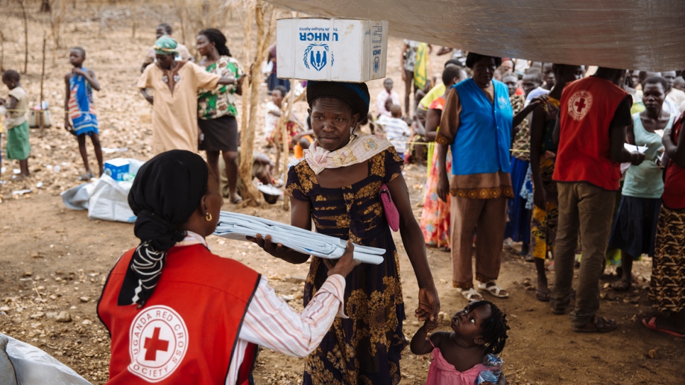 La réfugiée sud-soudanaise Opani Lilias, 28 ans, au centre, avec sa fille unique, Brenda, 21 mois, reçoit une bâche de la part d'un employé de la Société de la Croix-Rouge d'Ouganda, l'un des articles de secours de première nécessité fournis aux réfugiés à leur arrivée à l'installation d'Imvepi récemment établie, district d'Arua, Région du Nord, Ouganda. 