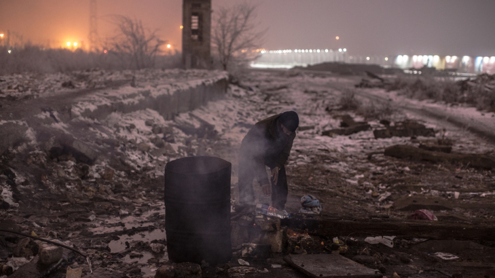 Un homme fait chauffer de l'eau sur un feu derrière un entrepôt. 