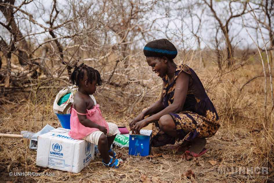 Opani unpacks a combined solar lamp and mobile phone charger as Brenda sits on a box of kitchen utensils, part of the Core Relief Items with which they were provided.