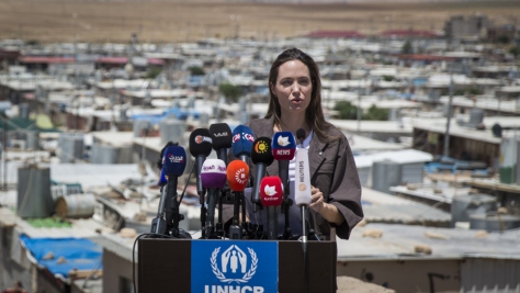 Angelina Jolie stands at a podium covered with news microphones, with Domiz refugee camp in the background
