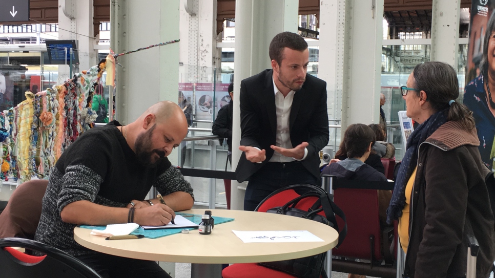 Seb Toussaint (centre), un artiste franco-anglais qui dessine des graffitis sur les murs de camp de réfugiés à travers le monde, parle à une voyageuse à la Gare de Lyon, le 24 octobre 2017, tandis qu'Omar Ibrahim (gauche), un artiste syrien, réfugié en France, dessine en calligraphie arabe le nom de la voyageuse, dans le cadre de la semaine de sensibilisation du HCR à la situation des réfugiés dans le monde. Omar Ibrahim est professeur de calligraphie à Science Po Paris. 