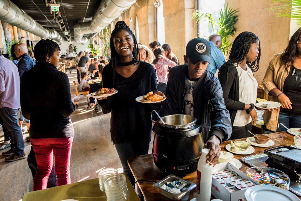 A women, carrying plates of food, smiles at the camera. Other people around her select food from a buffet table in a restaurant.