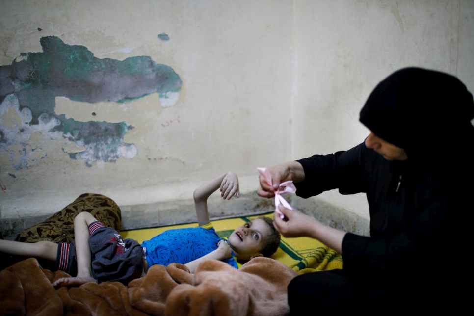 اللاجئة السورية غانية حموي تلعب مع ابنها عبد الهادي في منزلهما في مخيم الحسين الحضري للاجئين  في الأردن. غانية لديها ثلاثة أطفال يعانون من شلل دماغي ومن الصرع، وقد تلقت هنا رزمتين من الحلويات.