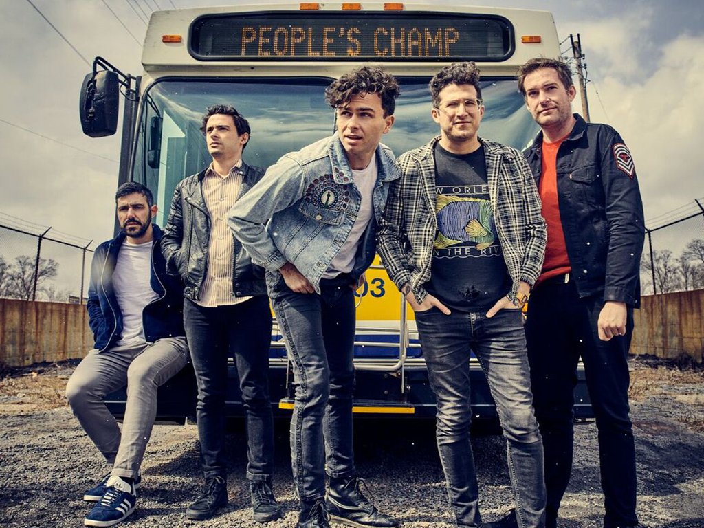 Max Kerman (centre) stands with fellow band members from Arkells in front of a yellow city bus bearing the sign "People's Champ."