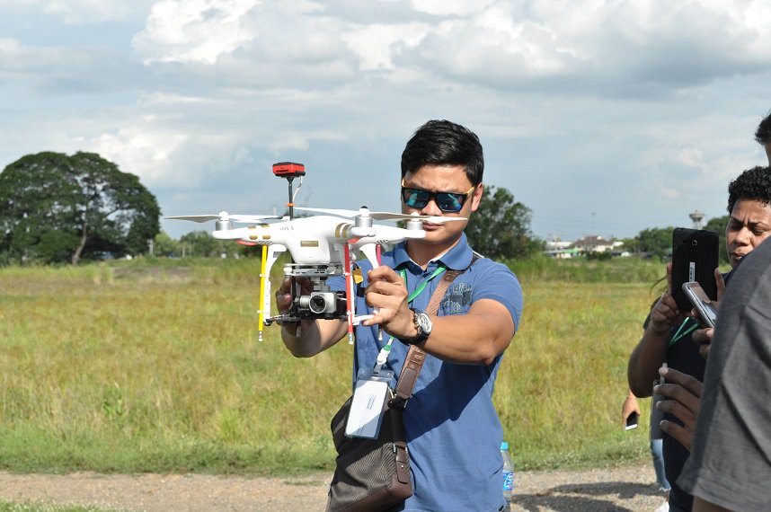 A trainer sets up a drone outside in the Rice Research Center, Thailand with participants recording through their mobiles