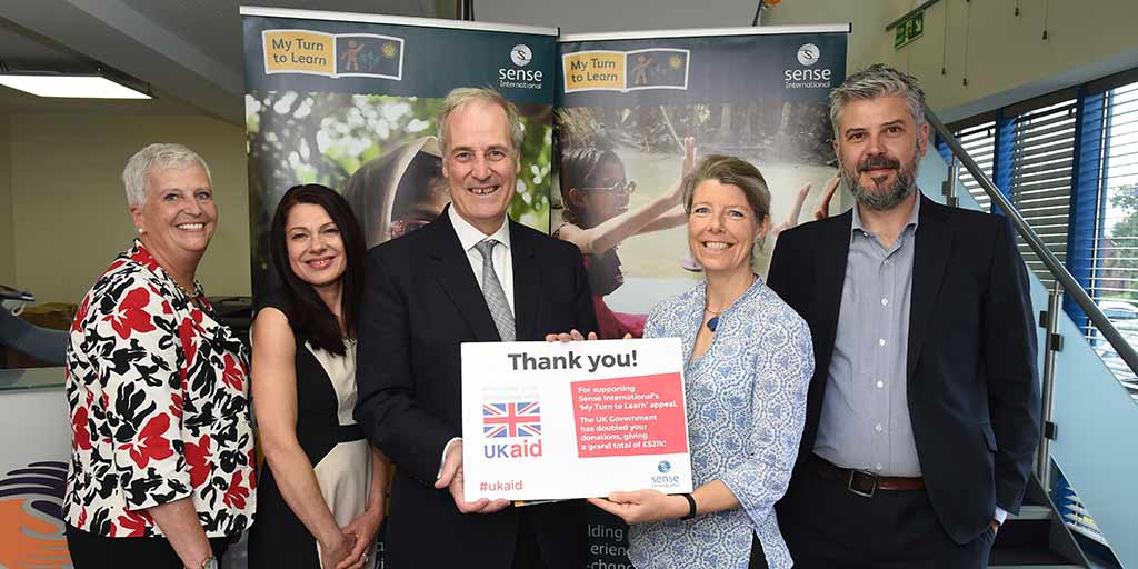 Two smiling men and three smiling women hold a sign that says "Thank you". Behind them a banner says "My Turn To Learn"