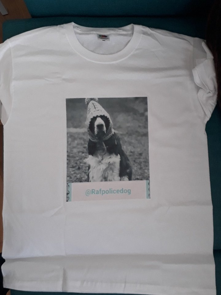 white round neck t-shirt with black and white photo in centre rectangle box.
of Daniel the Spaniel RAF Police Dog wearing his knitted hat with huge pom pom on top. tied under his chin. not sure of his back ground as that area is deliberately fuzzy.