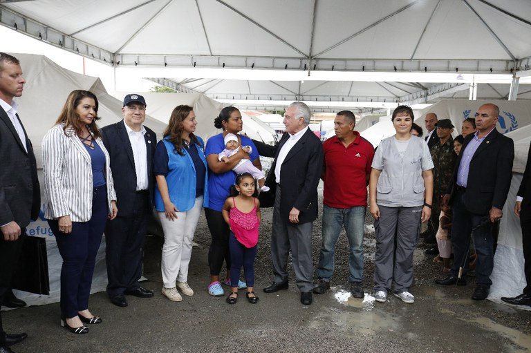 #PraCegoVer: Debaixo de uma tenda branca estão várias pessoas olhando para o presidente da República. No centro da imagem está o presidente da República, cumprimentado uma mulher que segura um recém-nascido.