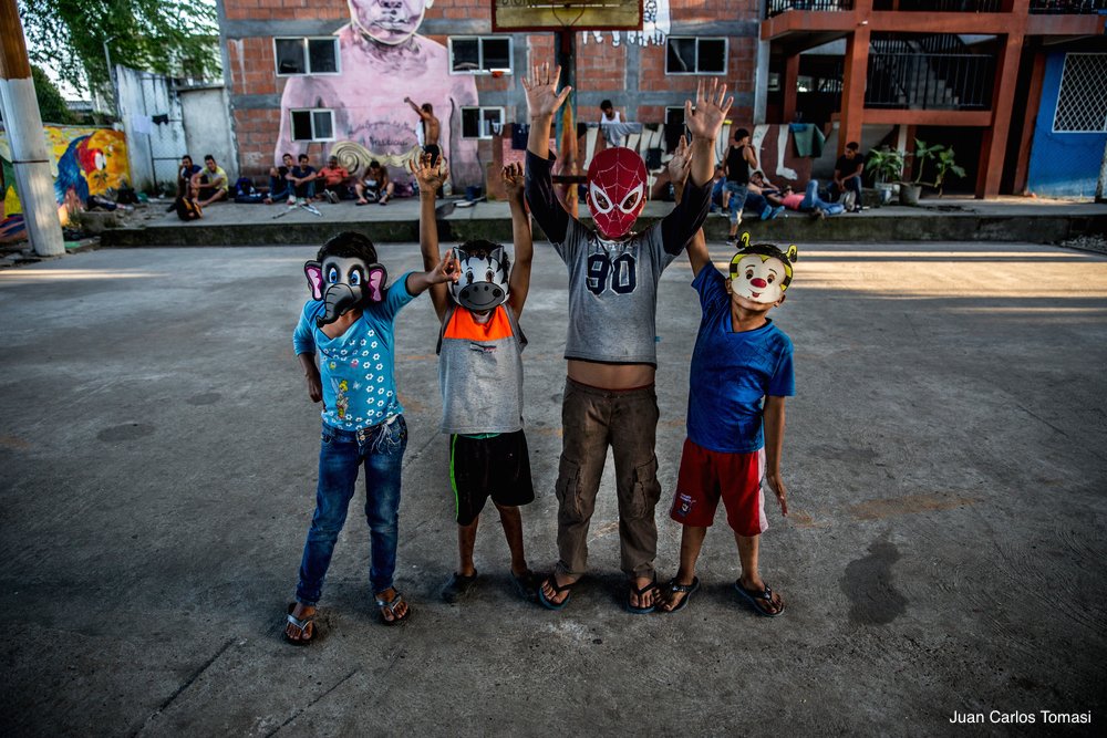 Four kids with animal and spiderman masks on raise their hands on a street in Mexico, with a shelter behind them. Many families flee Central America with their children because of violence in their countries