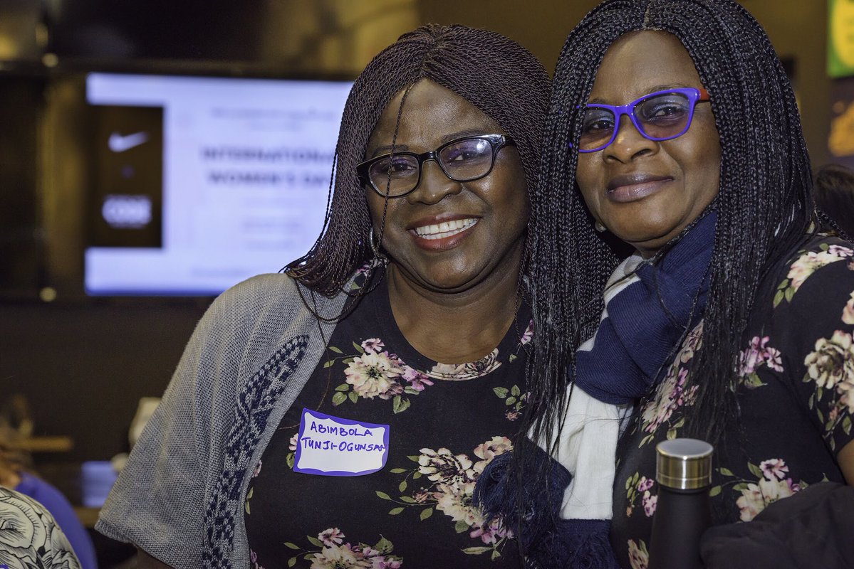 Two women in floral tops and glasses get together for a photo. A Nike projection screen in out of focus in the background.