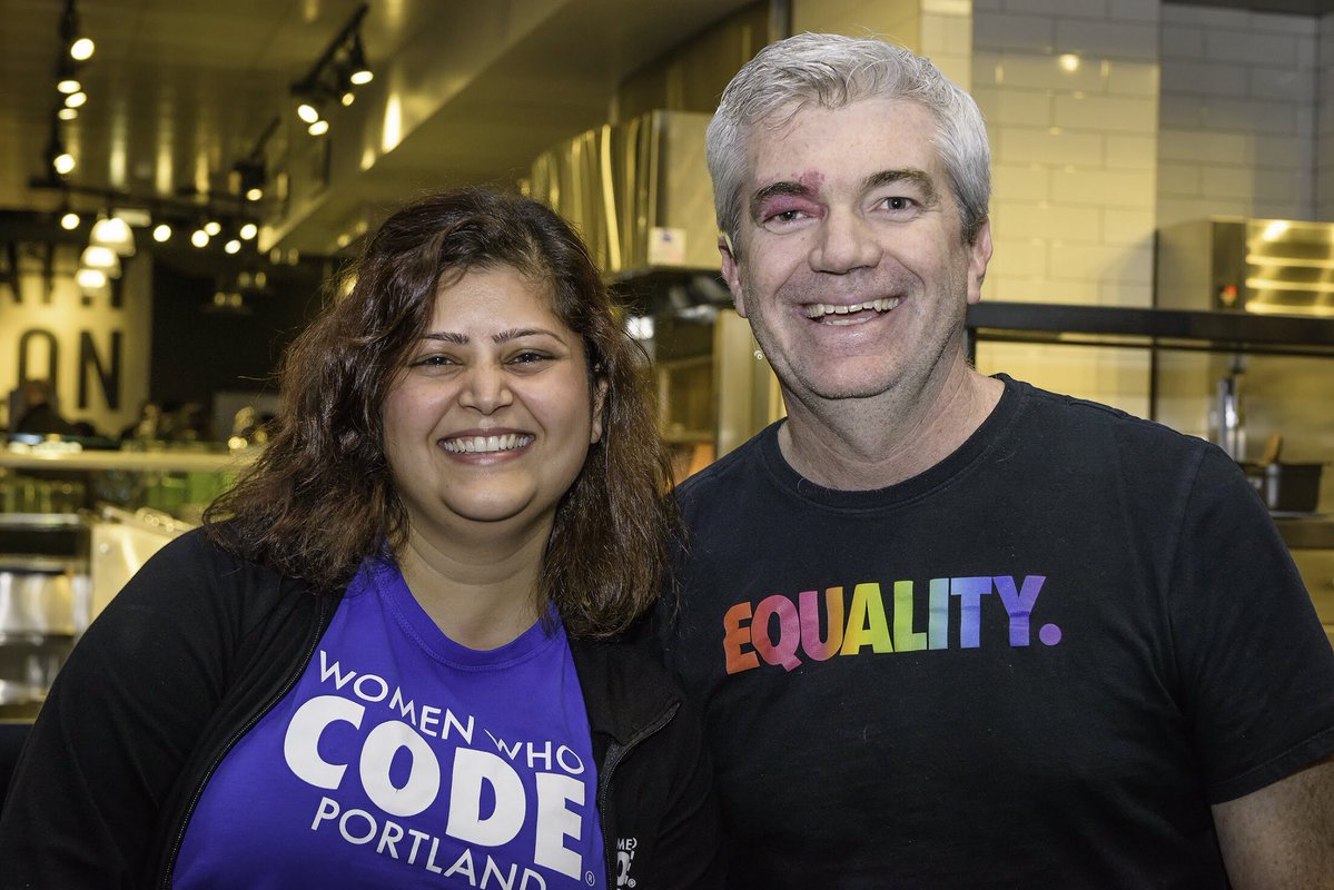 A woman and a man, Richa and Skip, get Tony for a photo. She is in her blue Women Who Code Portland T-shirt. He is in a black Nike, rainbow T-shirt.