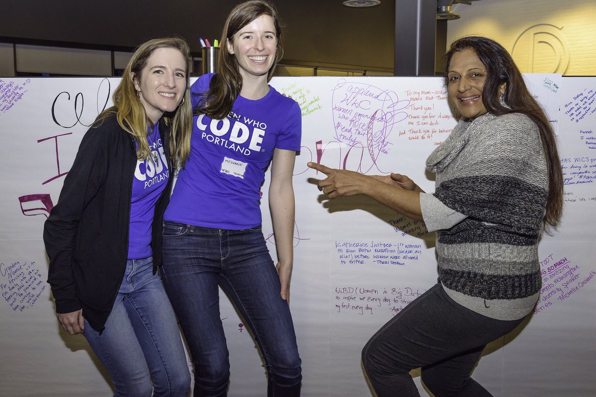 Two women in Women Who Code Portland Tees pose with another woman in a grey sweater, in front of the event’s #ApplaudHer wall.
