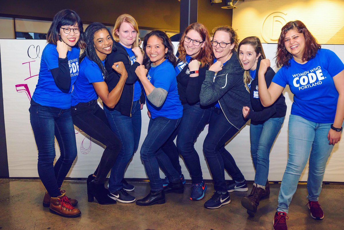 Eight women from the Women Who Code Portland leadership team, in their blue WWCode PDX T-shirts, pose in a bicep flex position, Rosie the Riveter style, in front of our #ApplaudHer wall.