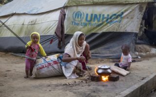 a woman sits on the ground watching a small silver pot cook on a small fire . she is outside and she is sitting on a sack with two children on either side of her. behind her is a tent branded with UNHCR