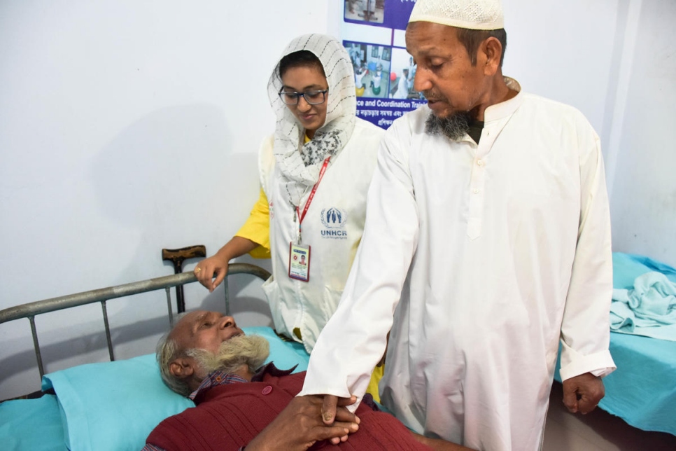a man in a white tunic and white hat shakes hand with a bearded man lying on a hospital bed wearing a red tunic, behind them stands a woman with a white headscarf and a yellow long sleeve shirt with a red lanyard around her neck holding an id badge and a white vest atop her yellow shirt with the unhcr logo on the chest