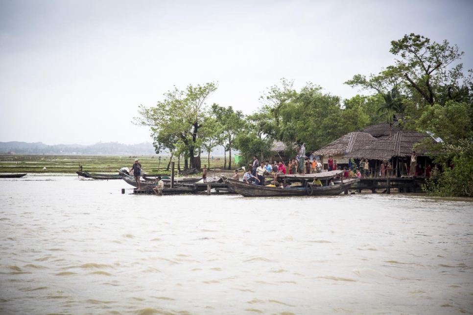 a photo from a river looking at two straw thatched huts with protruding docks coming out from in front of it and a few canoe like boats docked at the dock and people crowded around it