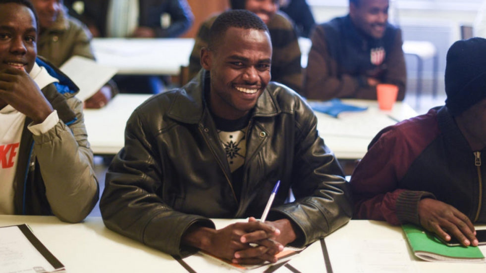 Man smiling at the camera in a class setting, there are other people sitting around him out of focus