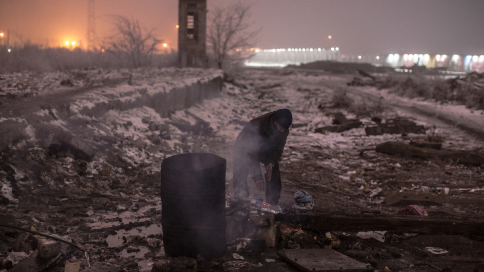 A man heats water on a fire behind a warehouse.