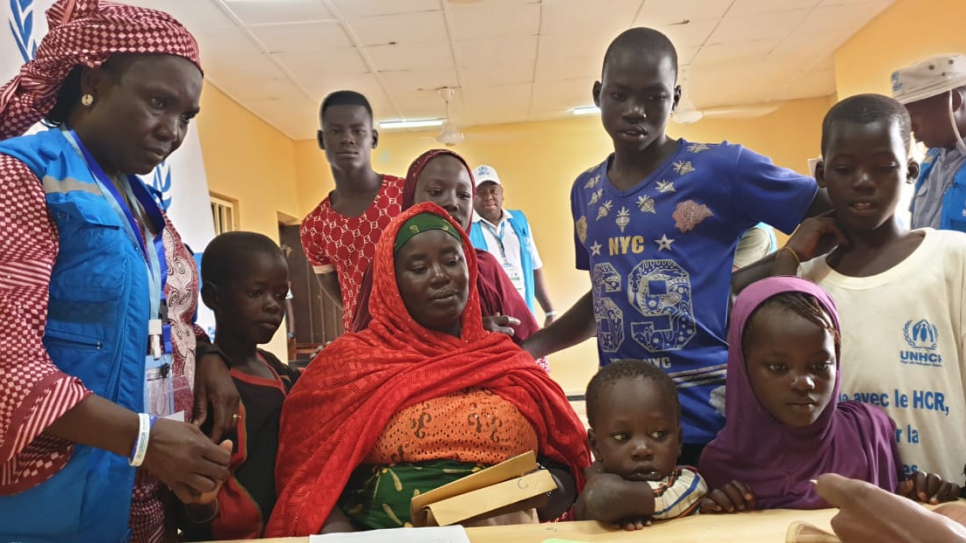 Jumai Mallum (seated) waits with her children as UNHCR staffer Veronica Yohanna (in blue vest) helps them through a registration process at Damare transit centre in Nigeria. 