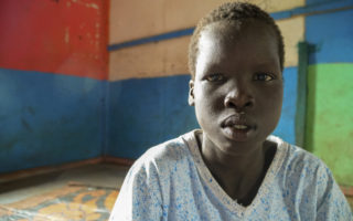 a young boy with a blue soccer jersey on looks directly at camera with multi coloured wall in the background