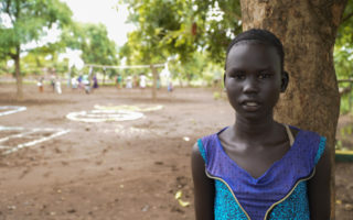 Young South Sudanese refugee girl poses in front of a camera in a blue and purple dress against the backdrop of an open field surrounded by trees