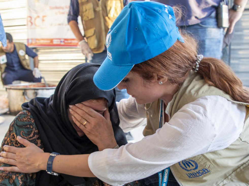 A woman from Syria cries beside another woman wearing UNHCR branded clothing.
