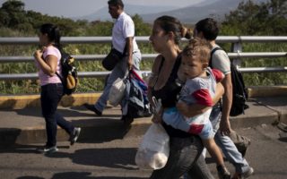 a group of people walk on what appears to be a bridge, closest to the camera is a woman carrying a plastic bag full of supplies and a child on her hip, she is wearing grey denim pants and a black tank top, her hair is tied back in a tight bun