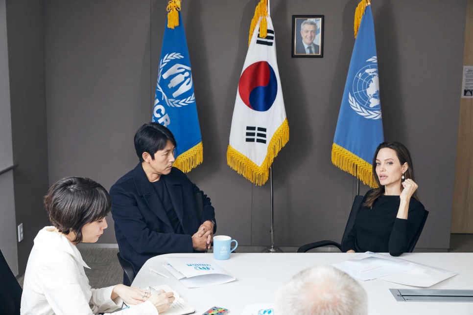 Special envoy Angelina Jolie and goodwill ambassador Jung Wuo-Sung sitting at a table in front of three flags, the two outer flags are UN flags and the middle flag is the Korean Flag. There is a woman sitting to the left of them also at the table.