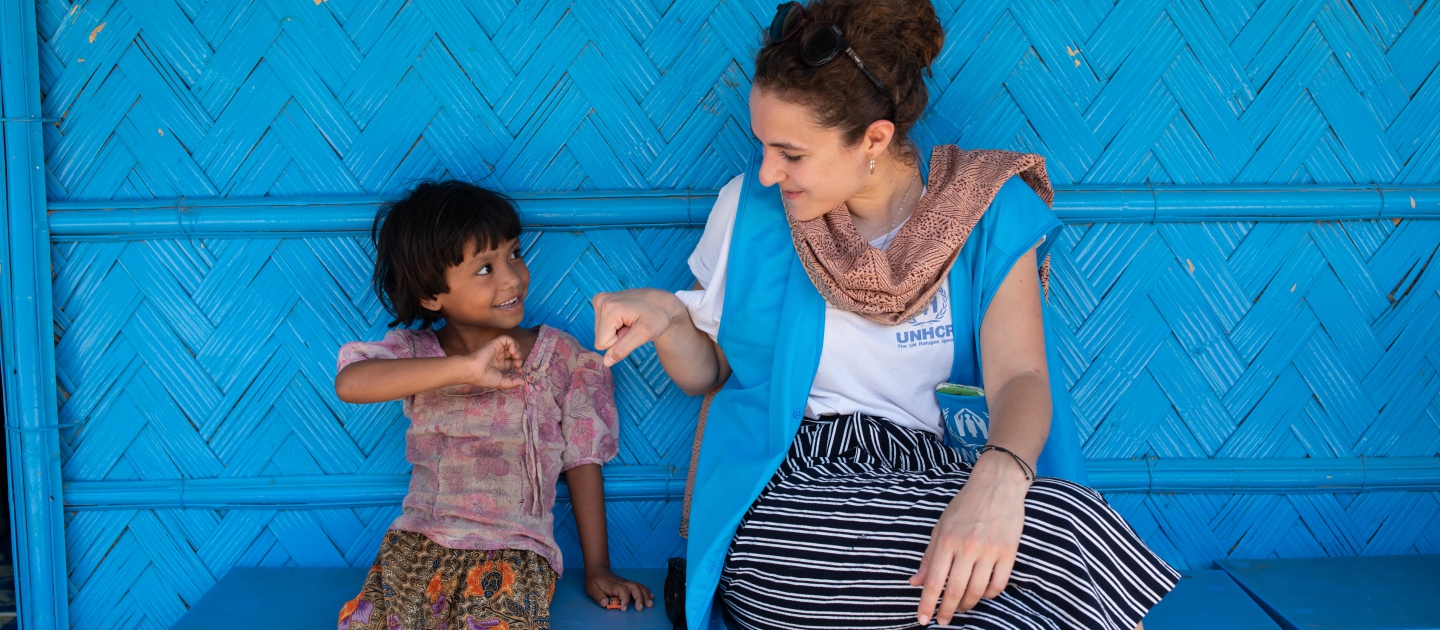 UNHCR FAQ Frequently asked questions - A young Rohingya girl sitting with a UNHCR staff member on a bench in front of a nutrition centre in a Cox'a Bazar refugee settlement.