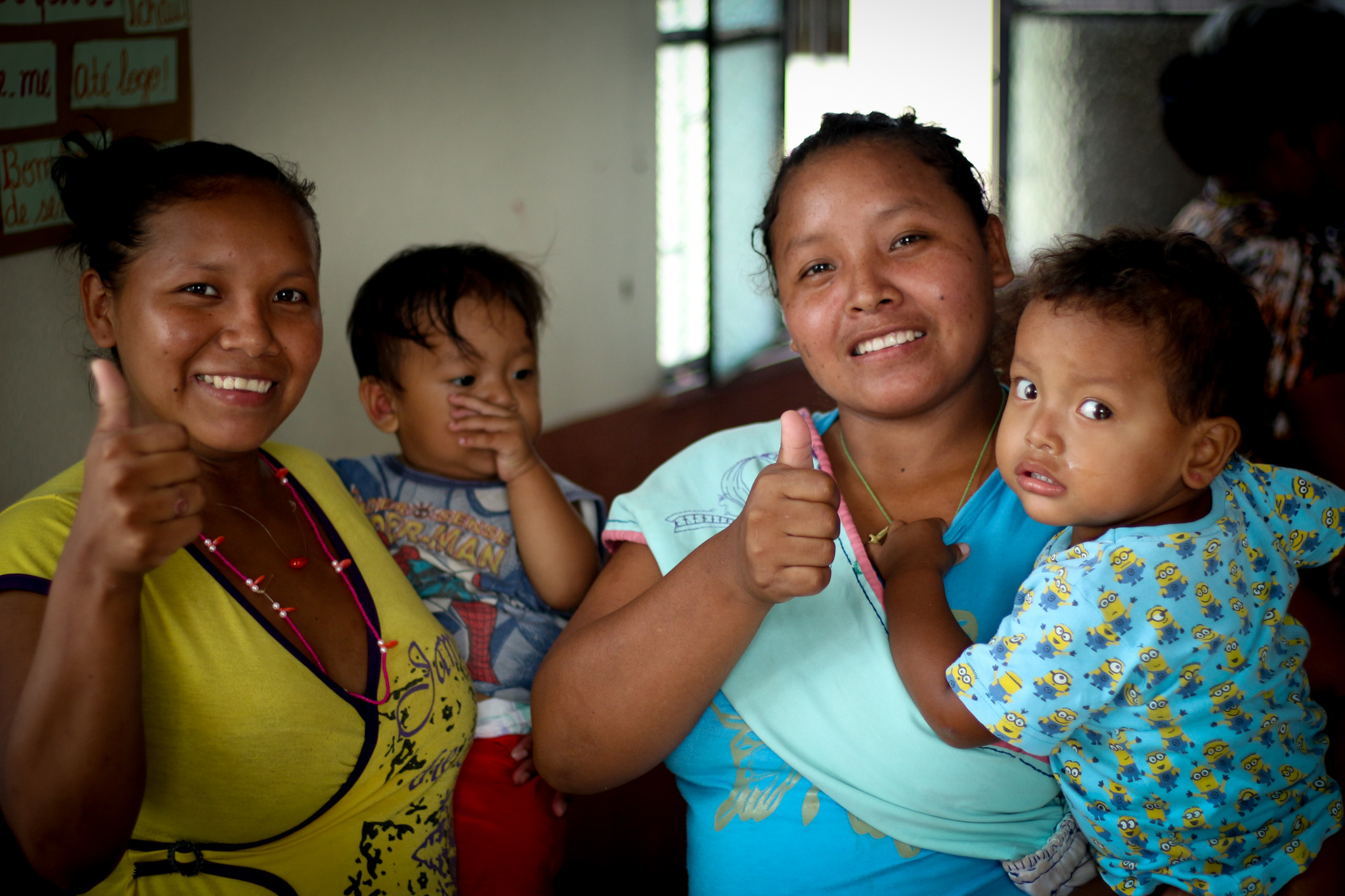 Two women smile facing the camera. They are both holding children.
