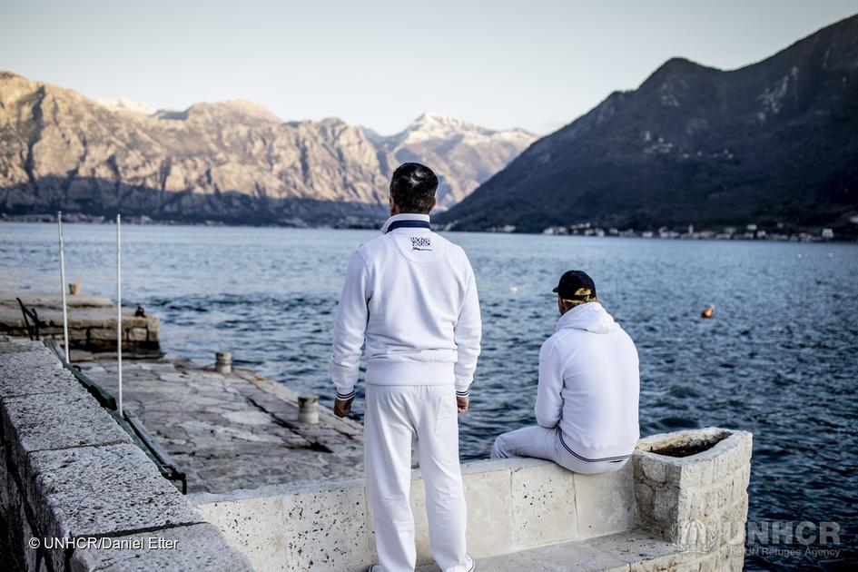 Two men sit with their backs to the camera. They are looking at a lake with mountains in the background