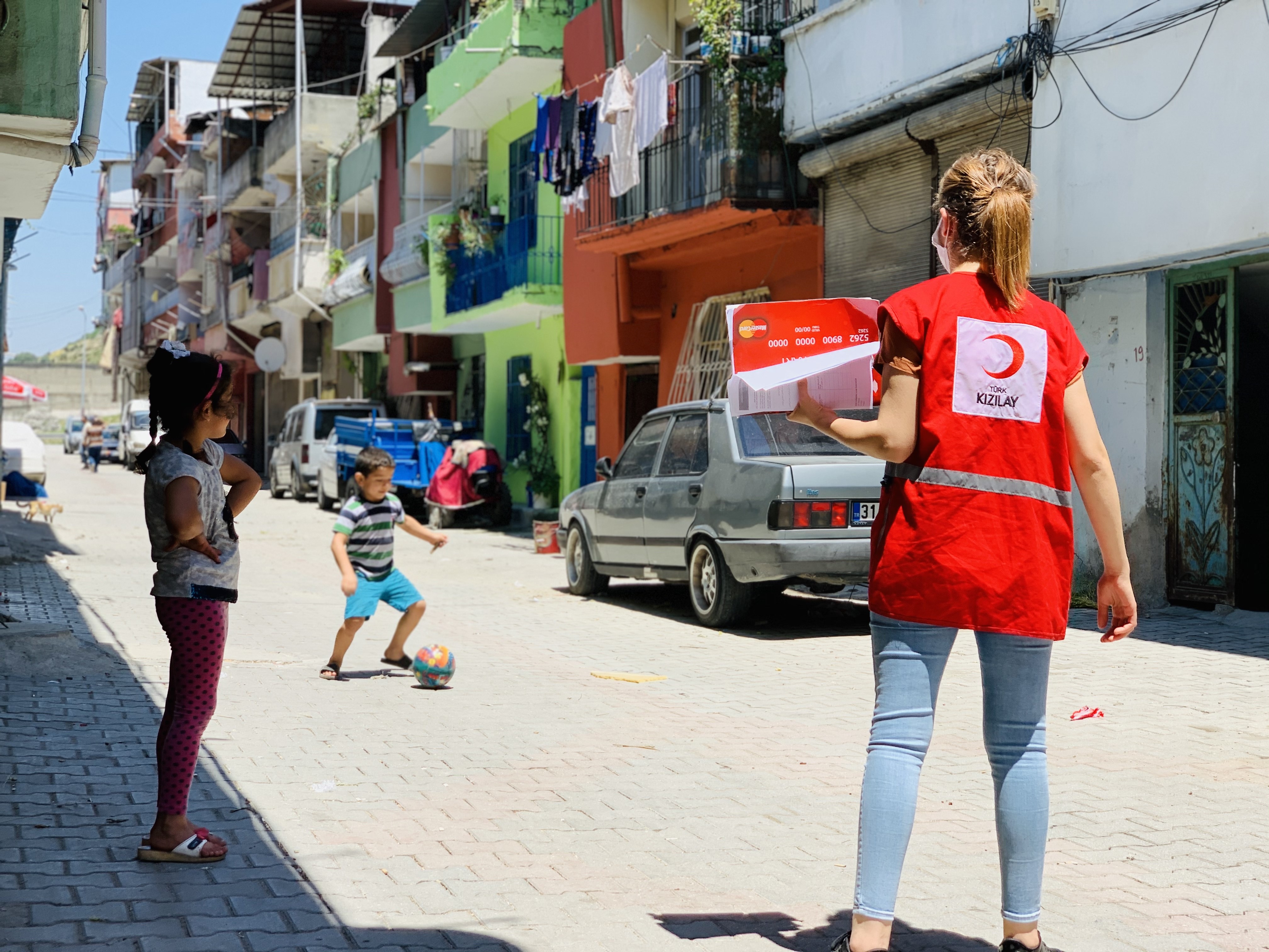Two children play ball in the street with a red crescent worker