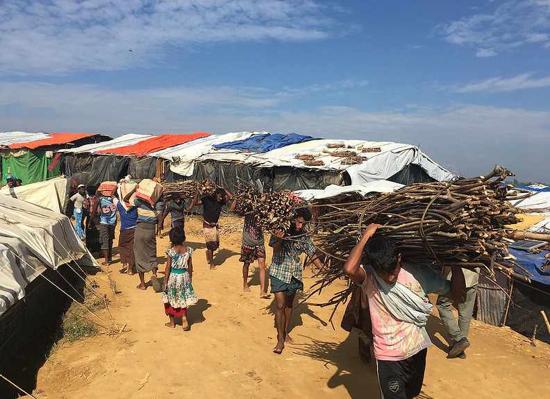 People carrying firewood over their shoulders in a camp setting. 