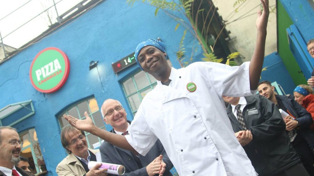 A chef standing with his hands up and smiling, while others clap. 