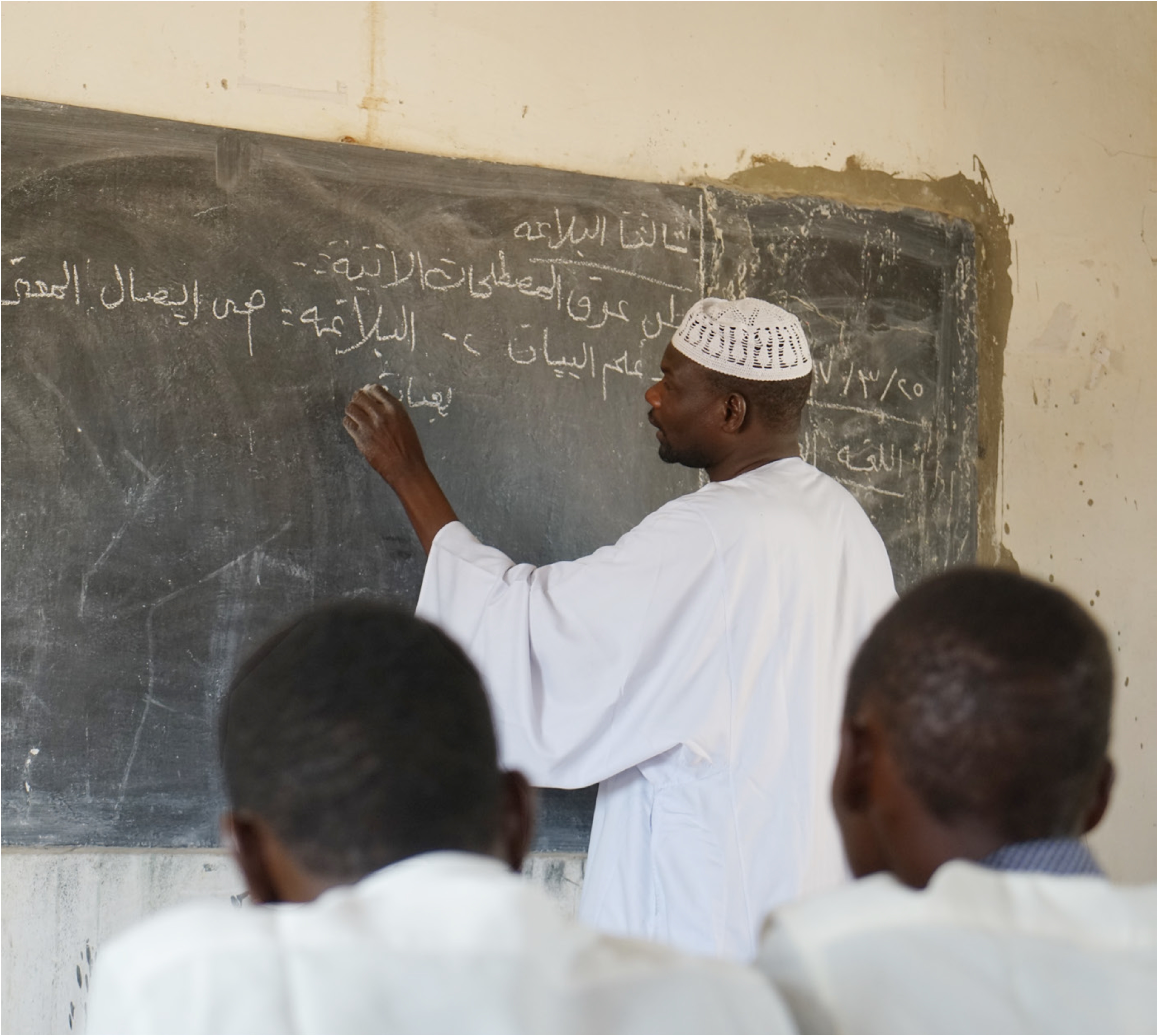 In the forefront to people sit back to the camera, in the background a man is writing on a blackboard