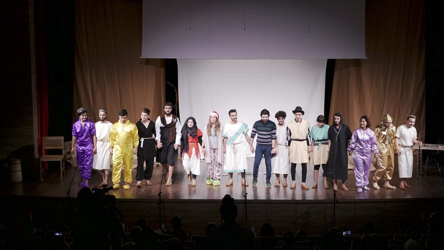 A big group of actors in costumes stand on stage, holding hands and facing the audience
