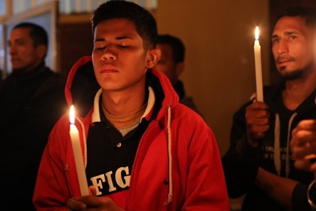 In the forefront, a man is holding a candle and has his eyes closed. In the background other people are holding candles. 