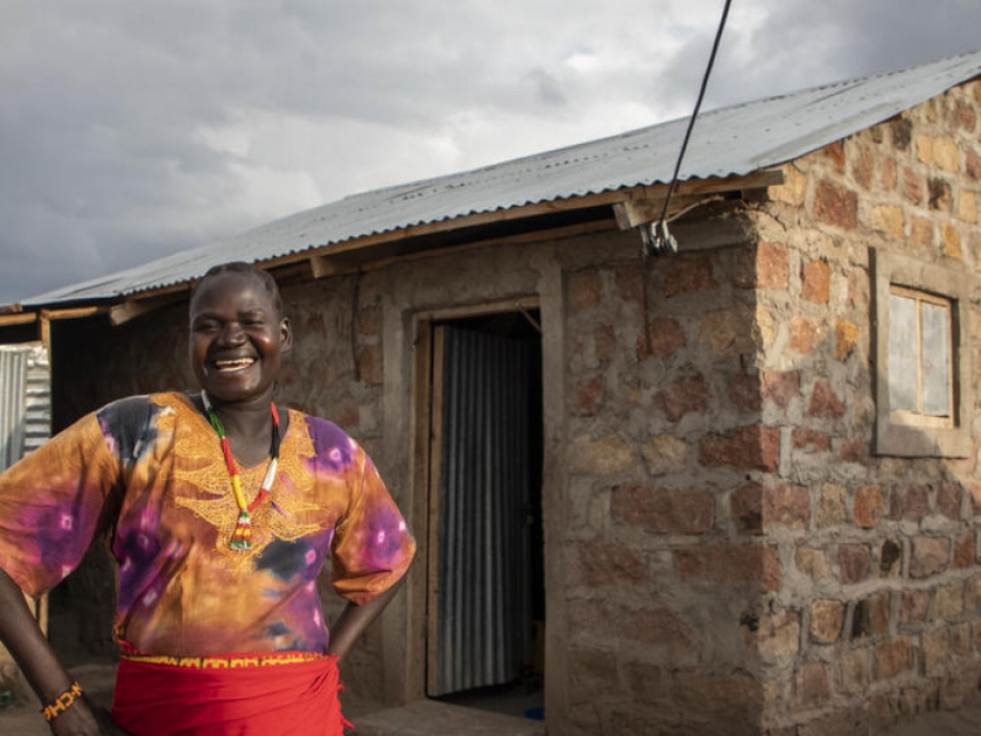 A smiling woman from Kenya stands outside of her house