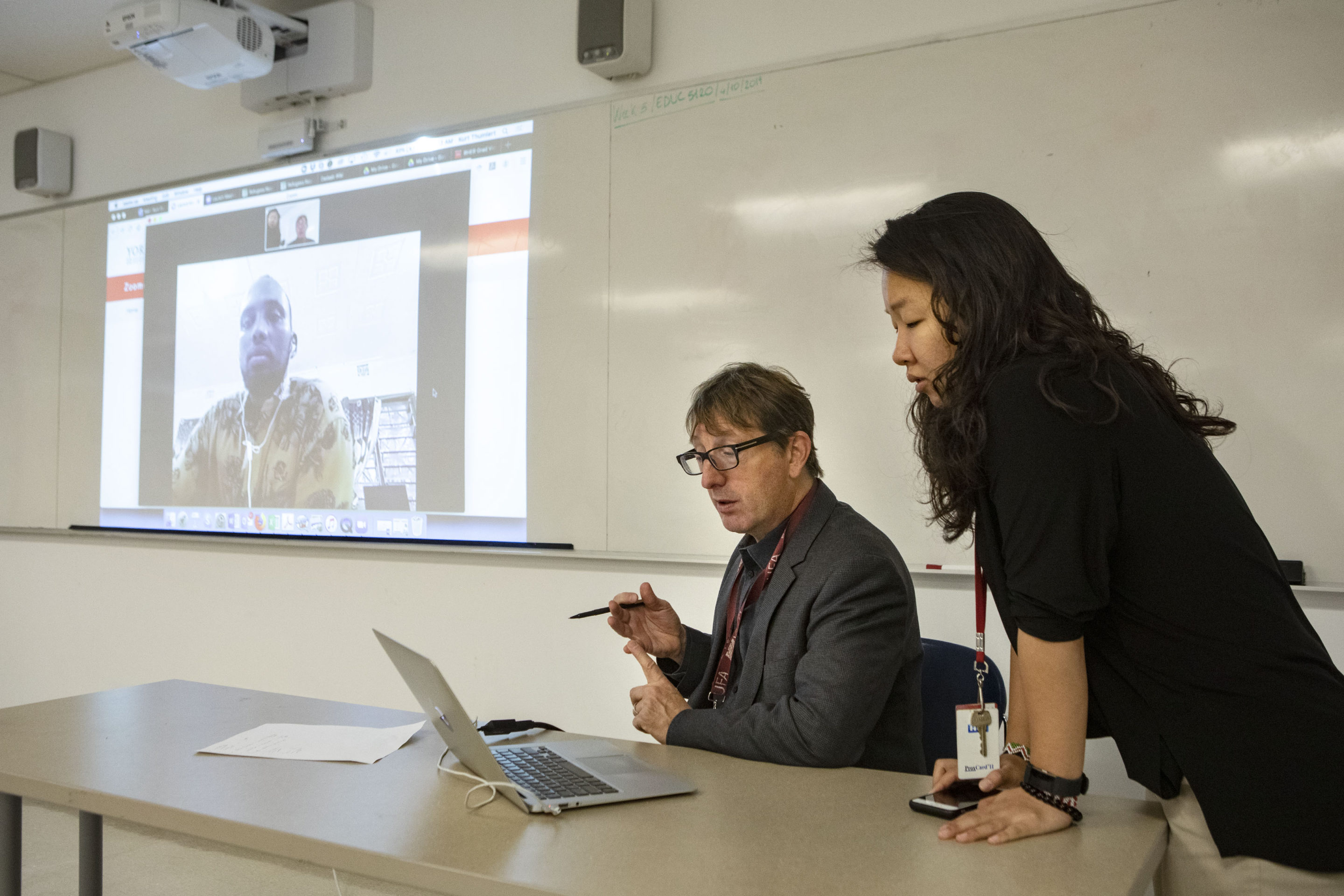 A man and a woman look at a lap top while a man is projected on a screen on the wall