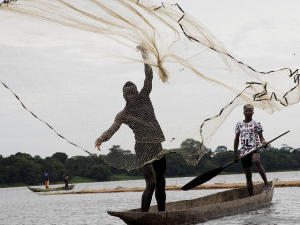 Two Central African men stand on a boat while one man throws a net in the water