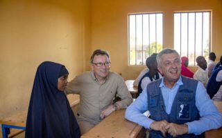 man in blue shirt and vest smiles looking at something behind a camera while he sits at a school desk with a woman in a black hijab and a man wearing a brown shirt, glasses and khaki pants