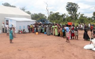 A group of Congolese people from DR Congo stand beside a shed with UNHCR branding