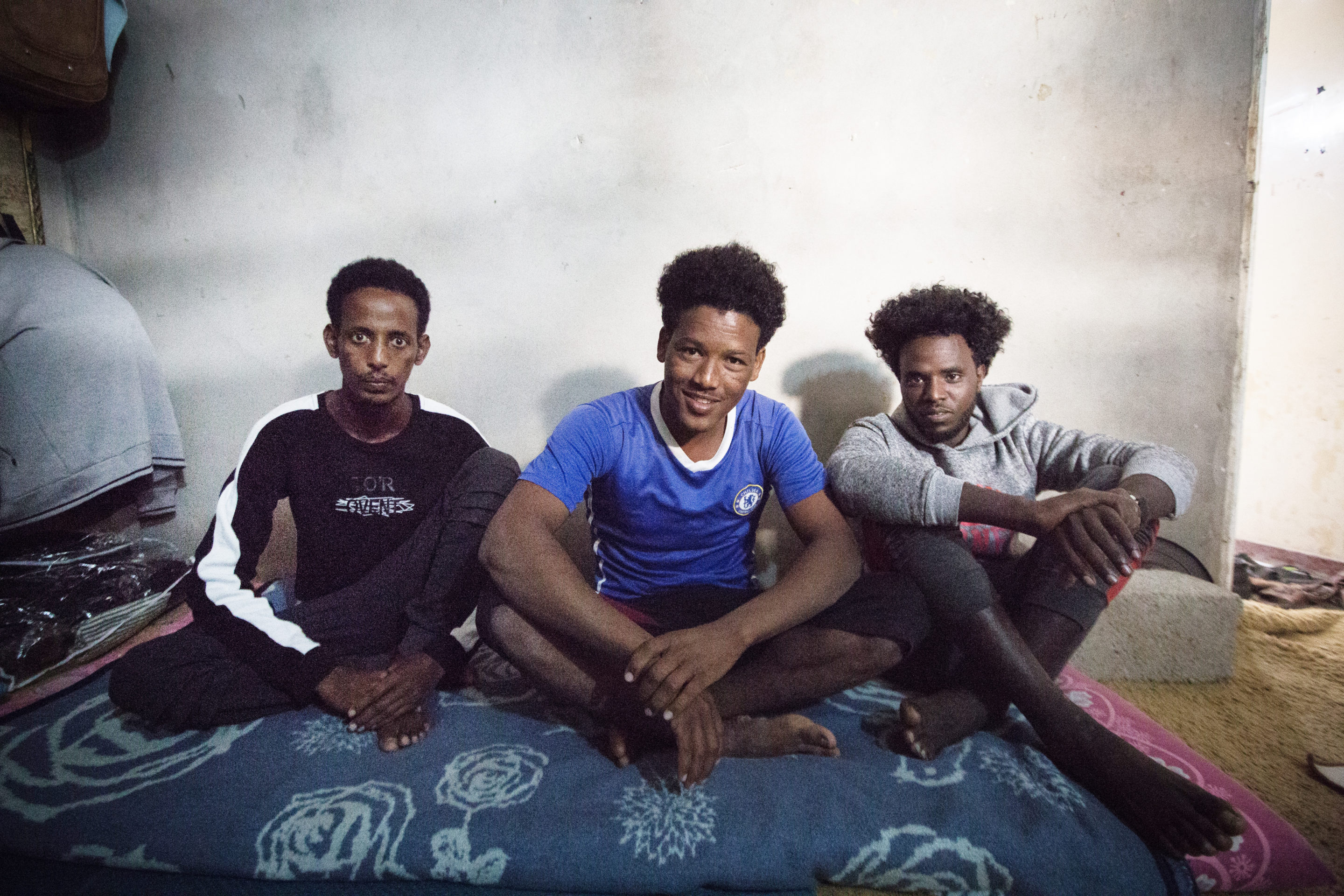 Three men sit side by side on the floor of their home