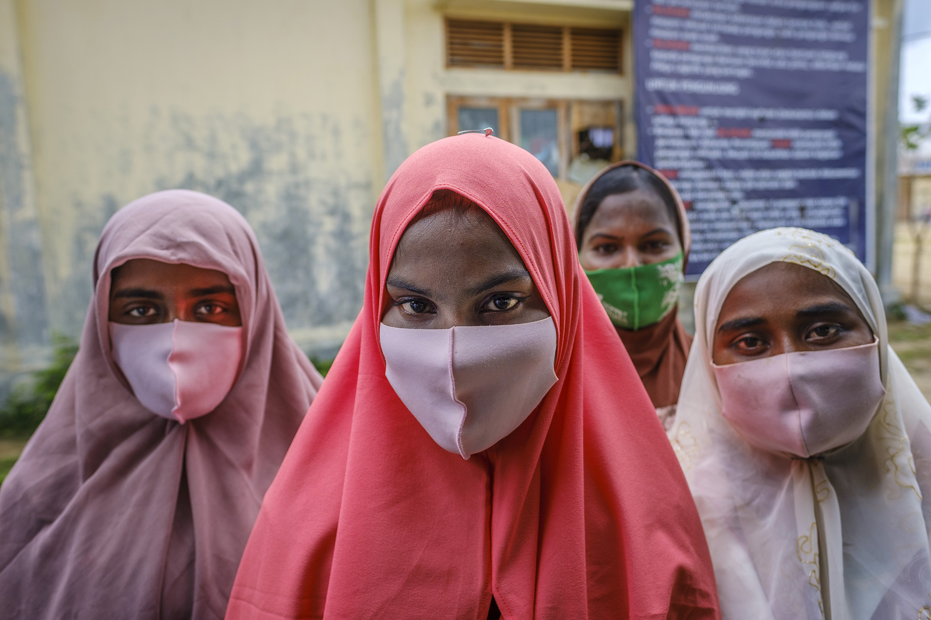 A female refugee wearing a salmon-colored shawl and a pink face mask looks at the camera, with a few fellow refugees in the background.