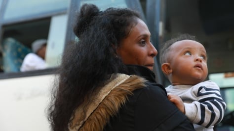 Solicitante de asilo lleva a su bebé en brazos mientras se prepara para abordar un autobús con dirección al aeropuerto, donde tomarán un vuelo para salir de Libia.